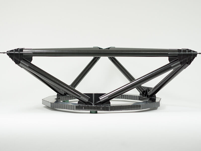 Image shows the completed TMT metrology fixture on a white surface and background. The structure is comprised of carbon fiber tubes attached at multiple points by dark metallic fittings. The bottom of the structure is a circular carbon fiber sandwich panel, with carbon plate covering the edges of the sandwich structure.  
